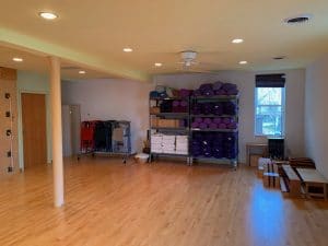 Yoga studio with hardwood floors, purple bolsters on storage shelves, folded blankets, and stacked chairs.