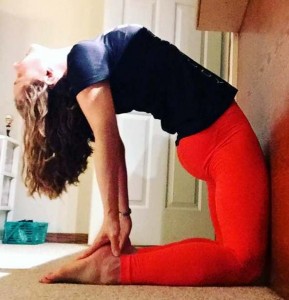 Woman in red leggings and blue top performing camel yoga pose (Ustrasana) on carpeted floor indoors.