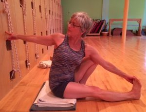 Older woman practicing seated yoga twist pose on mat in studio, holding rope on wall for support