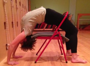 Woman performing a supported wheel pose backbend over a red folding chair with yoga mat on hardwood floor