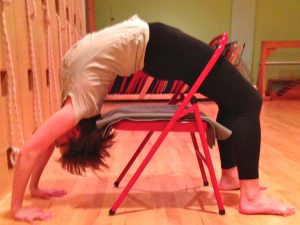 Woman performing supported wheel pose (Urdhva Dhanurasana) backbend over a red folding chair in yoga studio