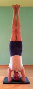 Woman performing a headstand yoga pose (Sirsasana) on a mat against a green wall, legs straight up.