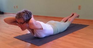 Woman performing yoga locust pose on a gray mat in a studio, lying prone with chest lifted and hands clasped behind head.