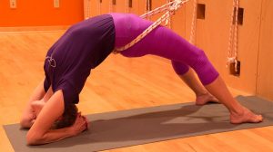 Woman in purple outfit performing yoga plow pose variation on gray mat in orange yoga studio with rope wall.
