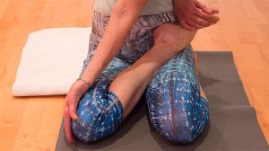 Person sitting on yoga mat in blue patterned leggings performing a seated foot and ankle stretch exercise.