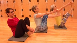 Three older women practicing boat pose (Navasana) on yoga mats in a wood-paneled studio with rope wall.