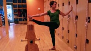 Woman in green top practicing yoga balance pose at wooden barre, holding leg extended forward in studio.