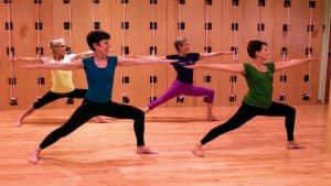 Four women practicing Warrior II yoga pose in a studio with wooden wall-mounted rope props.