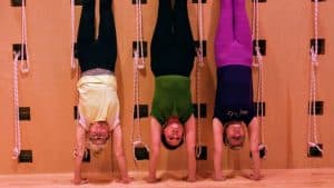 Three women doing supported headstands against a wooden Iyengar yoga wall with ropes in a studio class